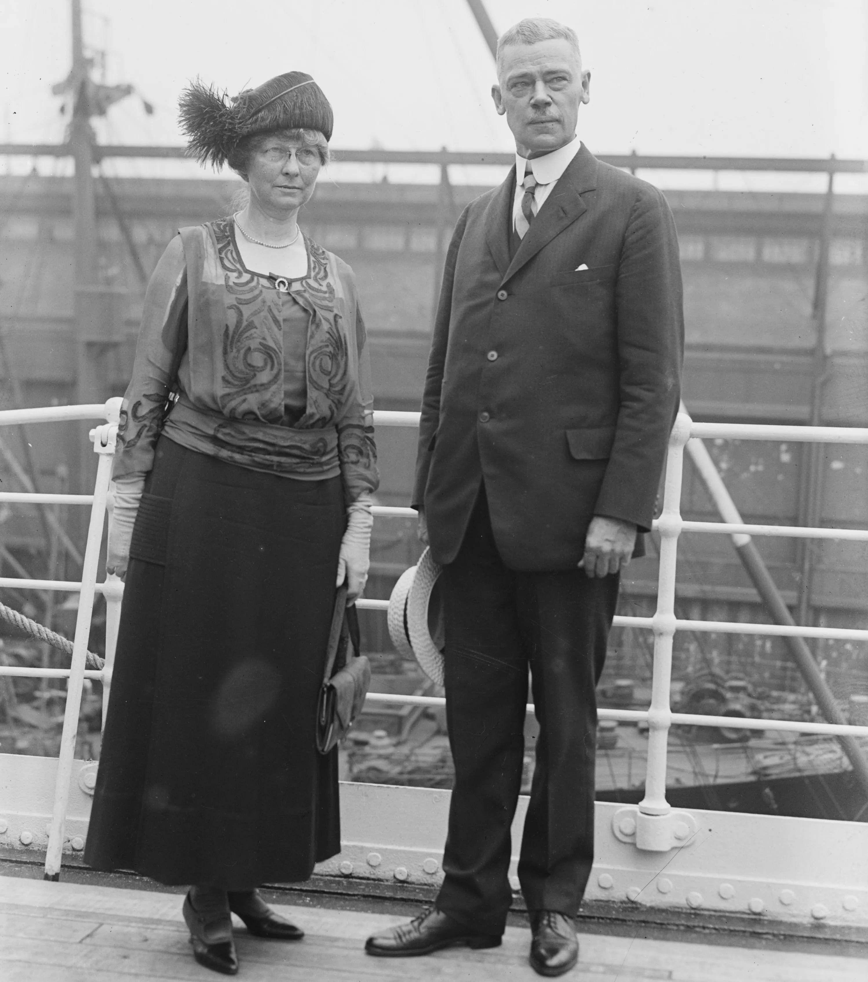 A black-and-white full-length photograph from the early 20th century featuring a man and a woman standing on the deck of a ship.The man, identified as Clinton Edgar Woods, stands on the right. He has short, light-colored hair and a mustache, wearing a dark three-piece suit with a high-collared white shirt and tie. He holds a light-colored boater hat in his right hand.The woman, his wife, stands on the left wearing a dark, floor-length skirt and a decorative, embroidered blouse with sheer sleeves. She wears a dark, wide-brimmed hat adorned with a large feather plume, spectacles, and light-colored gloves, holding a small handbag.They are positioned against a white ship railing, with the blurred industrial structures of a harbor and rigging visible in the background.