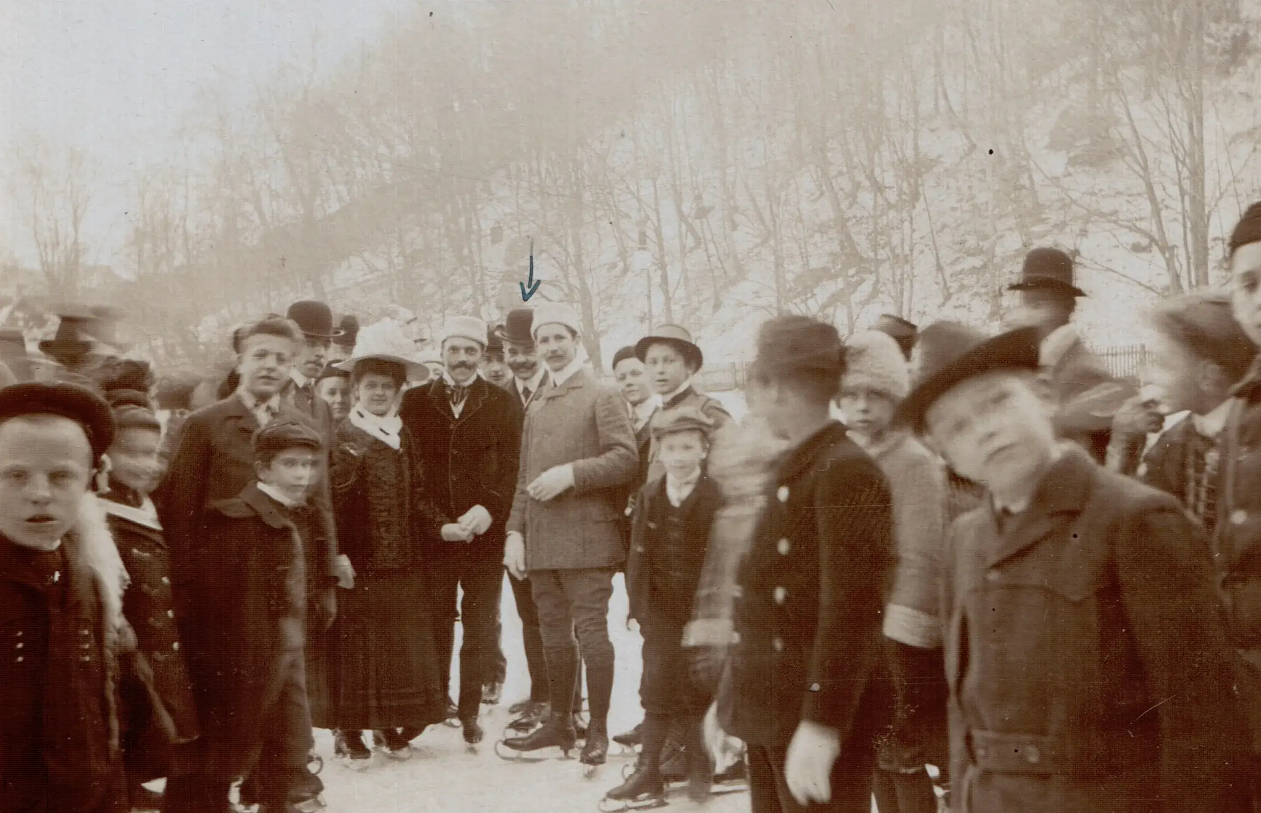 A sepia-toned historical photograph from 1909 in Graz, Austria, capturing a large group of people gathered on an ice rink. The crowd consists of men in formal coats and bowler hats, women in long skirts and ornate wide-brimmed hats, and several young children in winter caps. Many in the group are wearing antique ice skates, and the background shows a snow-covered hillside with bare trees.
