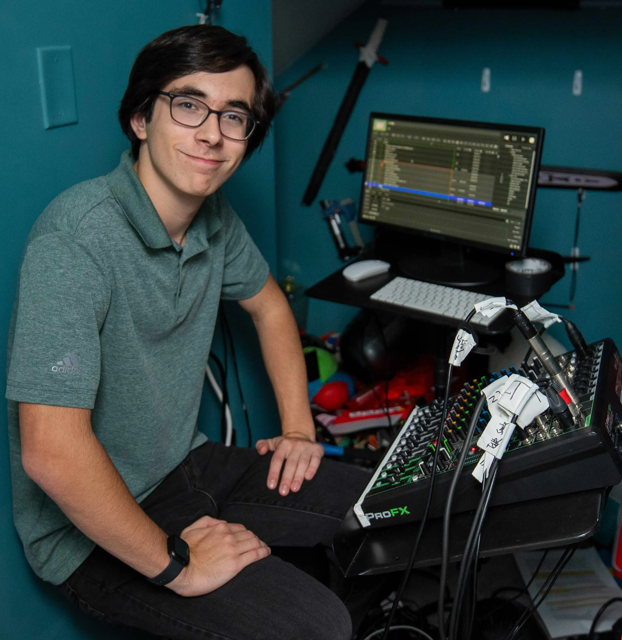 A young man with glasses and a green polo shirt sits in front of a technical workstation, featuring a computer monitor displaying audio editing software and a Mackie ProFX mixing console with labeled cables.