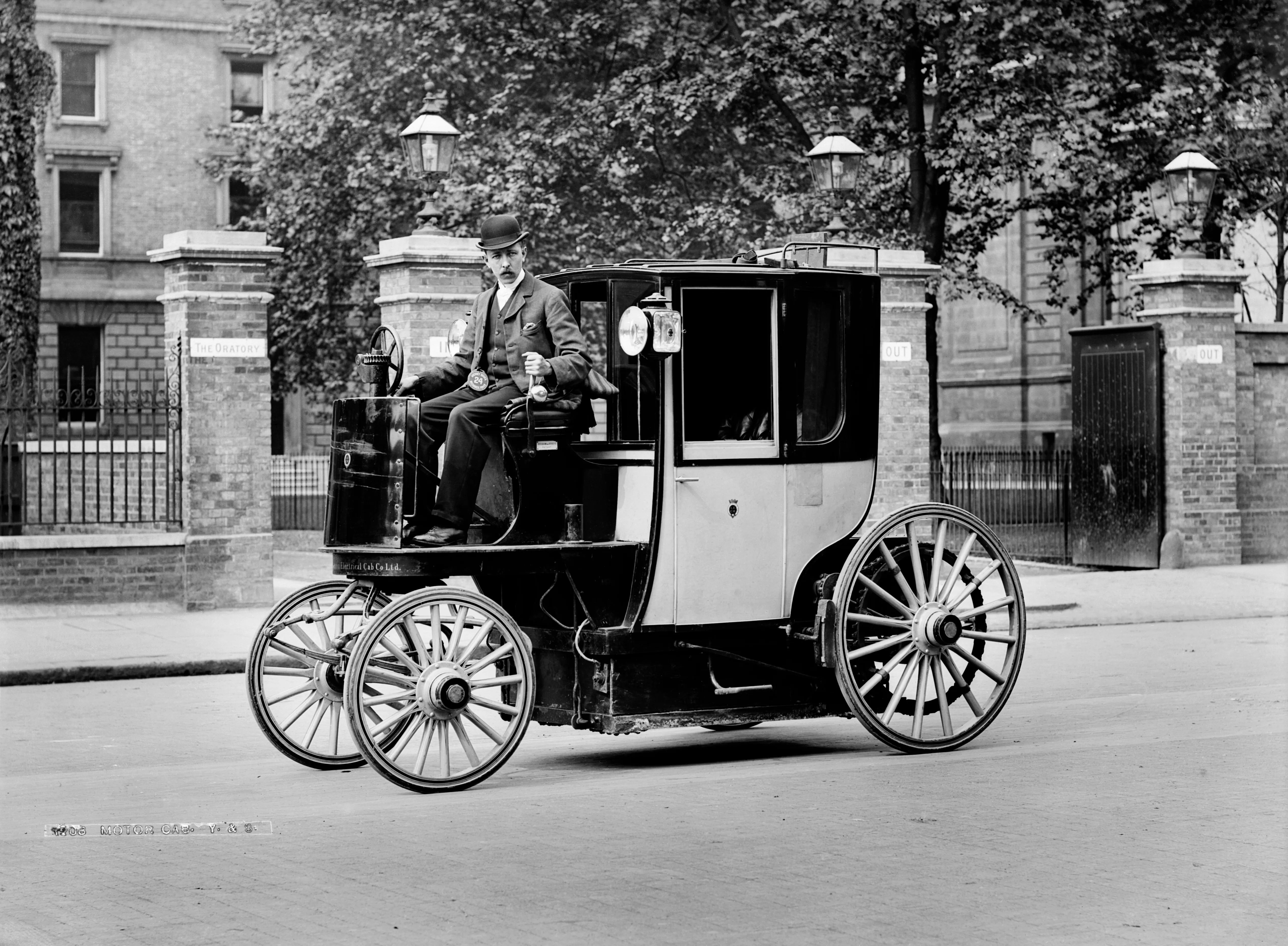 A black and white historical photograph of a Bersey Electric Cab, a vintage electric taxi, parked on a London street in front of the Brompton Oratory.The vehicle features a unique design with a large, enclosed passenger carriage at the rear and an open, elevated driver’s seat at the front. A man wearing a three-piece suit and a bowler hat sits at the steering wheel, which is a vertical column. The cab has four large, light-colored wooden-spoke wheels and two lanterns mounted near the passenger door. In the background, brick pillars with "IN" and "OUT" signs flank a gated entrance, and a large stone building is visible behind a row of trees.