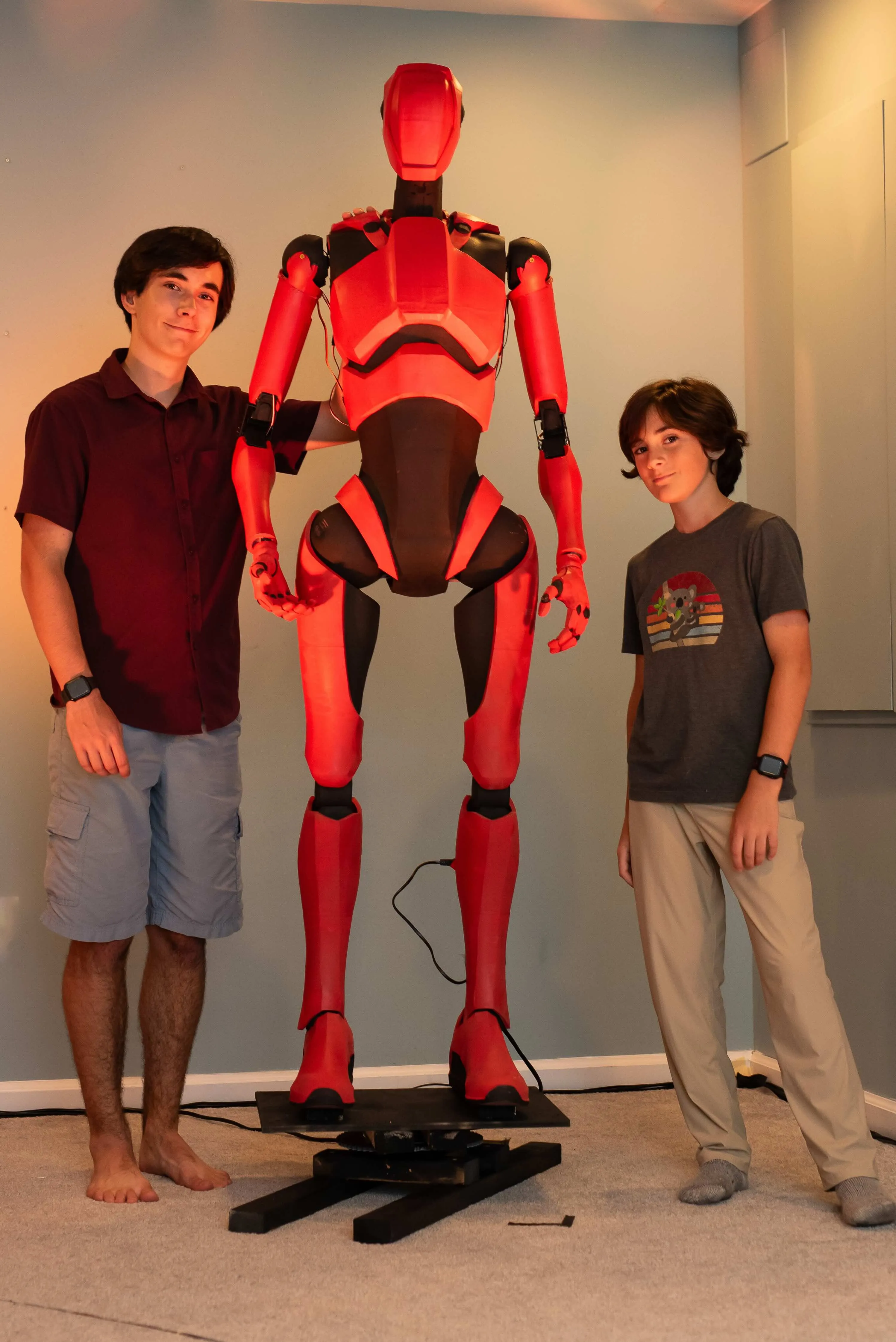 Two young men stand on either side of a tall, red and black humanoid robot prop. The robot is sleek and futuristic, mounted on a black platform in the center of a room with light-colored walls. The taller young man on the left wears a maroon button-down shirt and blue shorts, while the younger boy on the right wears a grey graphic t-shirt and tan pants.