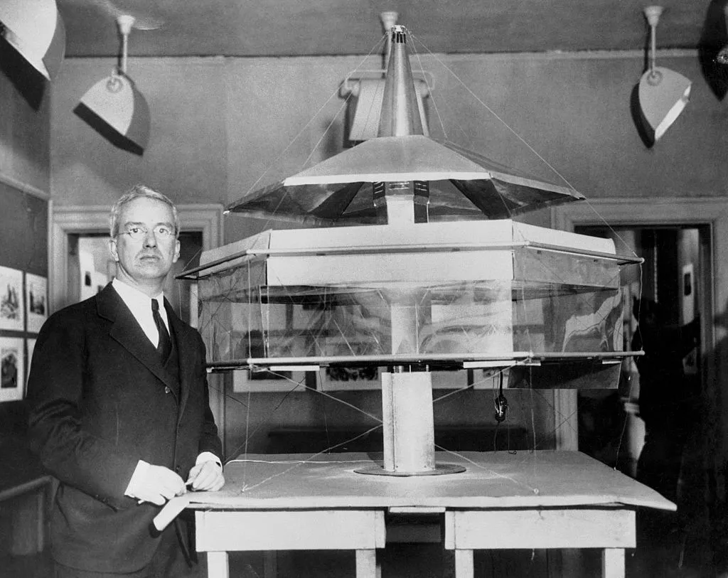 A man with gray hair and glasses in a suit stands next to a model of a futuristic flying saucer-like house.