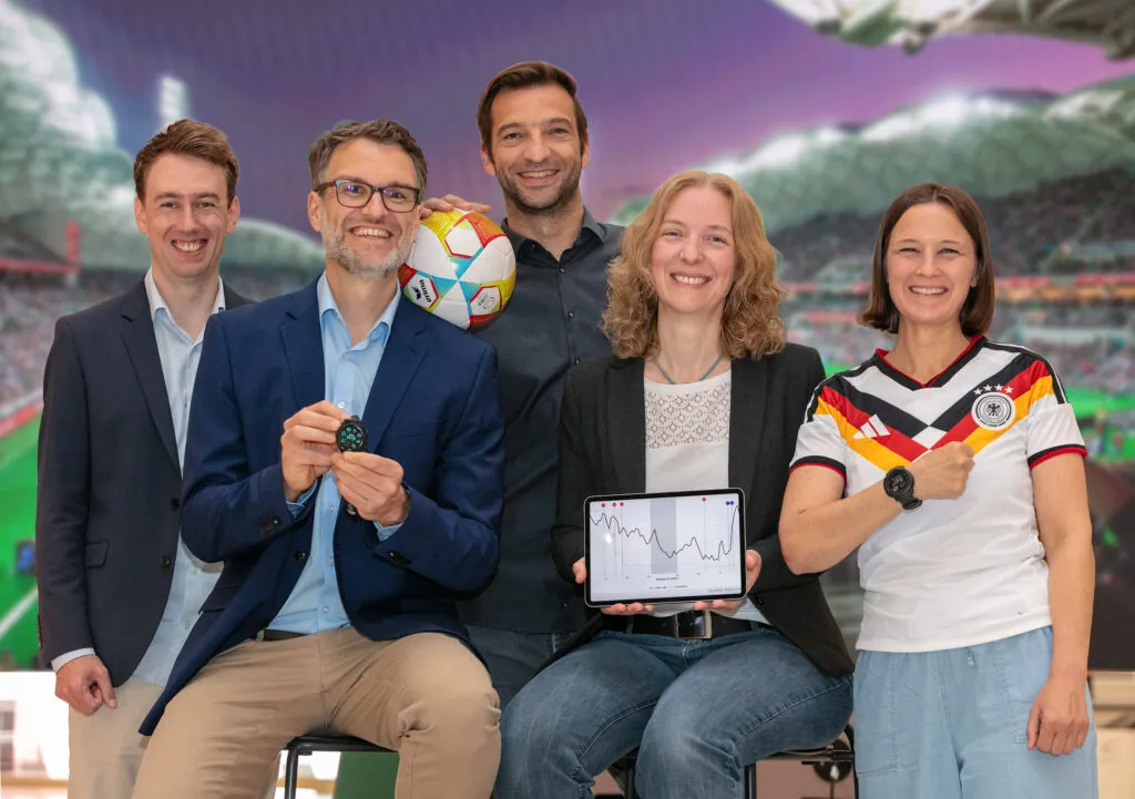 five scentists standing in front of a photo of a stadium. one holds a smartwatch, one holds a tablet with a graph, and one is wearing a german soccer jersey and smartwatch